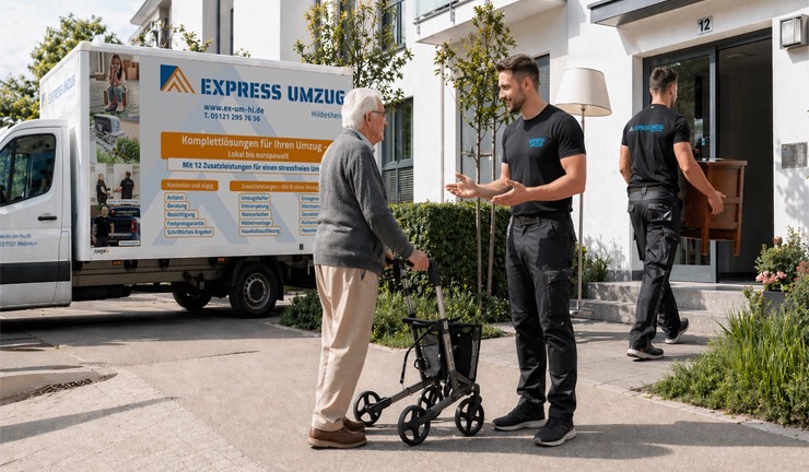Zwei Mitarbeiter von Express Umzug begleiten einen Seniorenumzug: Ein Mitarbeiter spricht vor dem Haus mit einem älteren Mann mit Rollator, während ein weiterer Mitarbeiter Möbel trägt. Im Hintergrund steht der Firmenwagen von Express Umzug.